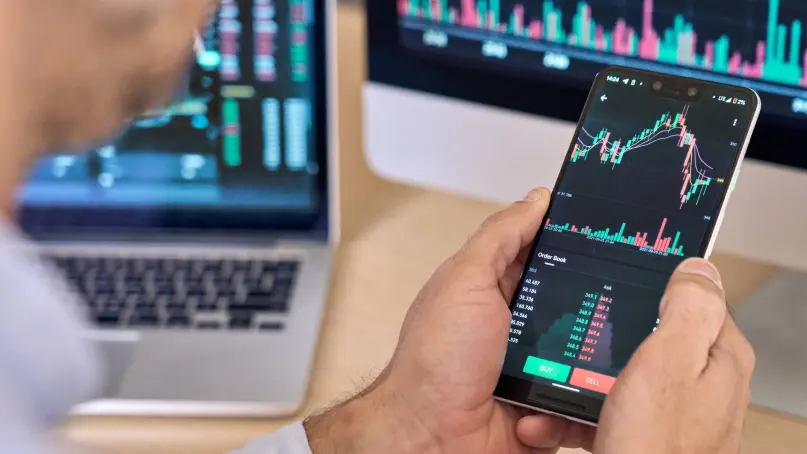 Das Bild zeigt eine Person, die ein Smartphone in der Hand hält, auf dessen Bildschirm ein Diagramm angezeigt wird, vermutlich zur Veränderung am Finanzmarkt oder an der Börse. Neben ihm befinden sich ein Computermonitor und ein Laptop, auf denen ebenfalls ähnliche Diagramme zu sehen sind. Insgesamt vermittelt das Bild eine Atmosphäre rund um Trading, Datenanalyse und Finanzen.